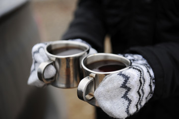 Fototapeta premium Two metal cups with coffee in hands in mittens close-up on car background. Brewing coffee in the open air