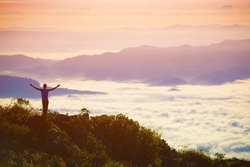 A man stands atop a mountain in nature with a relaxed posture. There is freedom with the atmosphere of the misty mountains, warm sunshine, forest trees in the morning, adventure travel on the holiday.