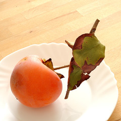 ripe kaki on a white dish, with green leaves, square.