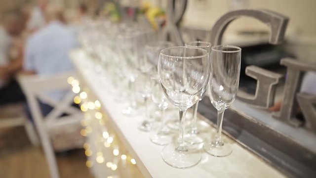 A lot of empty glasses on white table cloth. Yellow lights of a garland and unrecognizable people are sitting at the table on a blurred background.