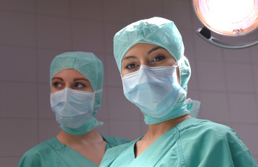 Two young women pose in a low lit operation theater.  Fully dressed as theater nurses with face masks  and green sterile medical work clothing.