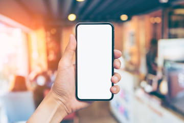 Man's hand shows mobile smartphone with white screen in vertical position, Blurred or Defocus image of Coffee Shop or Cafeteria for use as Background vintage tone. - mockup template and clipping path