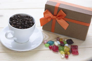 Festive still life with a brown gift box, cup of coffee with a saucer and spilled colored candies. The cup is filled with coffee beans. Close up. Light wooden background. Copy space