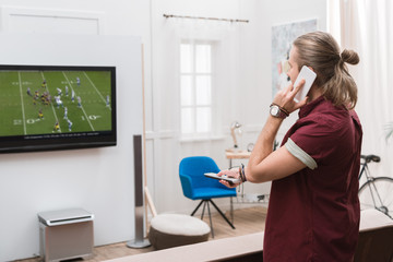 man talking on smartphone while watching football match at home