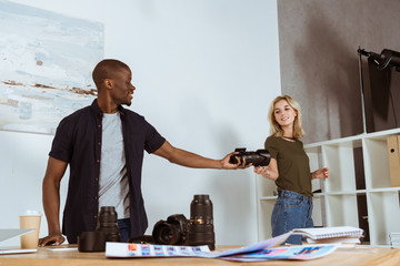 smiling caucasian photographer giving photo camera to african american colleague at workplace in...