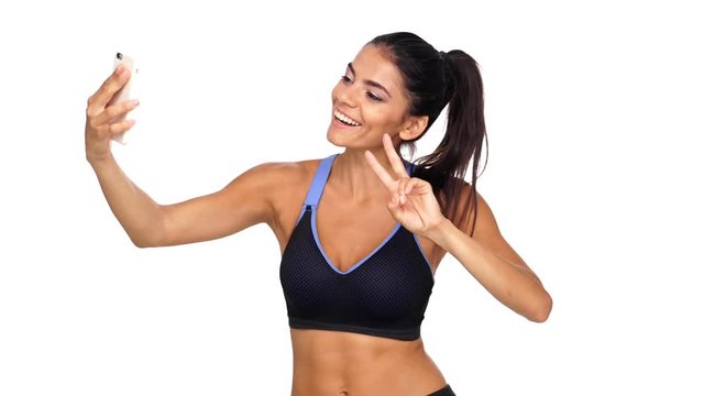 Smiling brunette sports woman making selfie while showing peace sign and her bicep over white background