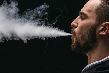 Young man vaping e-cigarette with smoke on black