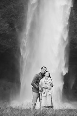 Iceland couple embrace near the big waterfall Wall of the water on the background. Woman wear beautiful linen dress and knitted scarf They are enjoy each other and nature around.Black and white photo.