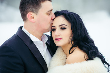 Horizontal portrait of the groom kissing the beautiful serious brunette bride into forehead. Winter time.