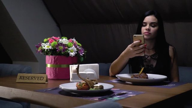 A woman sits at a reserved table in a restaurant near a bouquet and takes pictures of smart dishes on the smartphone