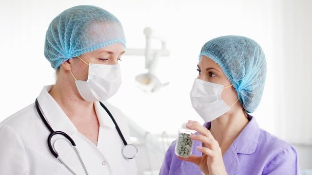 Two pretty female doctor colleagues in medical masks and caps discussing new experimental treatment at modern hospital room. Holding bottle of pills. Medical professional staff at work