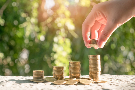 Hand putting money gold coins to coin stacks on blurred tree and sunlight background. Concept for Save Money, Financial, Business Growth, Investment