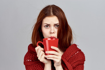 Red-haired girl on a light background in a red scarf drinks a hot drink from a mug