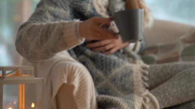 Cozy winter lifestyle. Young happy woman drinking cup of coffee wearing knitted sweater sitting home by the big window with winter snow tree background. Slow motion