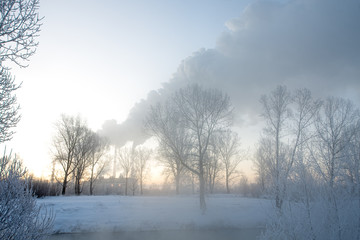 Snowy frozen landscape on sunrise with trees and smoke of factory
