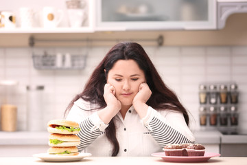 Overweight young woman with unhealthy food at table in kitchen