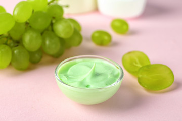 Bowl of body cream with grape on color background