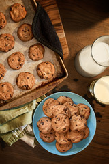Overhead view of cookie sheet and plate of chocolate chip cookies warm from the oven with glass of cold milk on vintage wood table