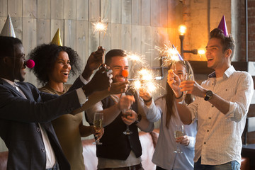 Happy multiracial people in party hats clown noses holding sparklers and champagne glasses celebrating New year eve together, excited diverse young friends having fun enjoying celebration together
