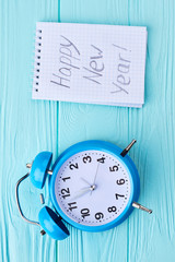 New Year decoration on wooden background. Blue alarm clock and paper notebook with message Happy New Year on blue wooden background, top view.