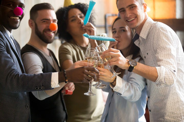 Multiracial young people in party hats clinking glasses celebrating New years day eve or birthday, diverse friends looking at camera having fun holding champagne, blowing whistles at celebration
