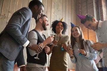 Multi-ethnic party people enjoying celebration together, having fun holding champagne glasses, multiracial diverse young friends celebrating joking laughing wearing funny hats standing indoors
