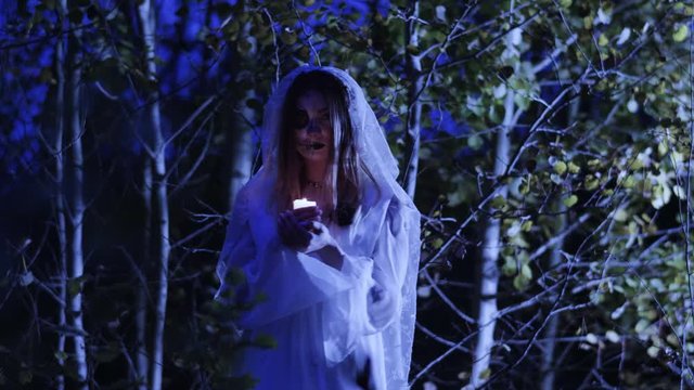 Halloween make-up. The young woman holding a burning wax candle in the arms in the dark forest at night. 4K