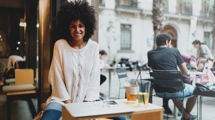 Portrait of a cheerful international student female with black curly hair wearing stylish clothes looking at the camera while sitting in a modern city coffee shop on a weekend day.