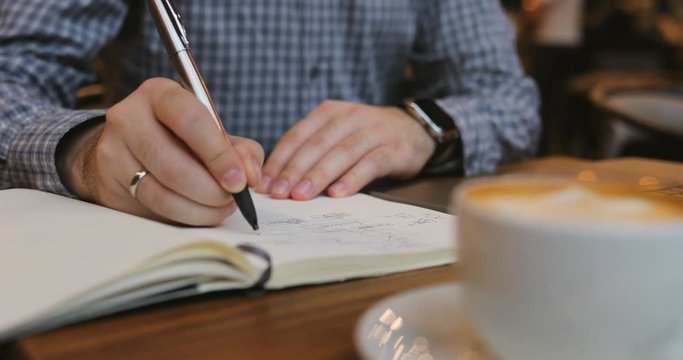 Close-up of a man's hand writes in a notebook in the foreground of coffee in a cafe break businessman code business cup