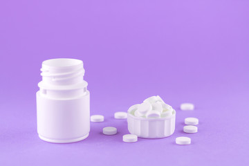 Close up white pill bottle with spilled out pills and capsules in cap on purple background with copy space. Focus on foreground, soft bokeh. Pharmacy drugstore concept