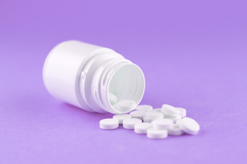Close up white pill bottle with spilled out pills on purple background with copy space. Focus on foreground, soft bokeh. Pharmacy drugstore concept