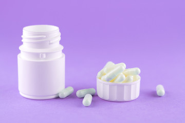 Close up white pill bottle with spilled out pills and capsules in cap on purple background with copy space. Focus on foreground, soft bokeh. Pharmacy drugstore concept