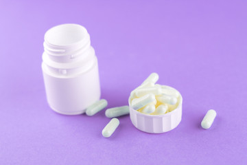Close up white pill bottle with spilled out pills and capsules in cap on purple background with copy space. Focus on foreground, soft bokeh. Pharmacy drugstore concept