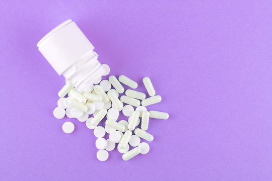 Close up white pill bottle with spilled out pills and capsules on purple background with copy space. Focus on foreground, soft bokeh. Pharmacy drugstore concept. Top view