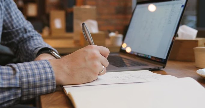Close-up of a man's hand writes in a notebook and use laptop in a cafe break businessman code business cup screen social media sitting