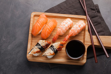A set of sushi with salmon, shrimp and eel with a bowl of soy sauce and wooden chopsticks on a wooden Board. Delicious Japanese food on a dark stone background. Top view