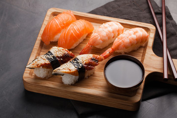 A set of sushi with salmon, shrimp and eel with a bowl of soy sauce and wooden chopsticks on a wooden Board. Delicious Japanese food on a dark stone background. Top view