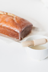 Pastry brush with cup of lemon sugar icing to coat the freshly baked and homemade lemon pound cake or Madeira cake with lemon sugar icing on white table