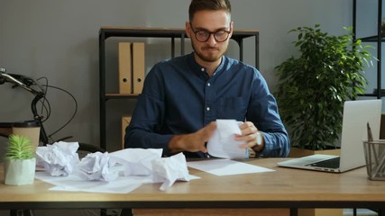 Portrait of angry man in the glasses trying to write a business plan than screwing up paper and throwing it to the table in the office. - Powered by Adobe