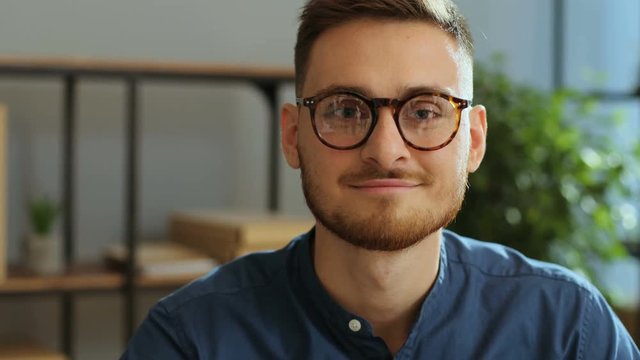 Portrait of successful young businessman in the glasses looking straight at the camera and smiling on the grey office background.