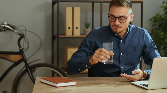 Young sick business man in the stylish glasses taking pill during work time in the modern office. Taking medecine
