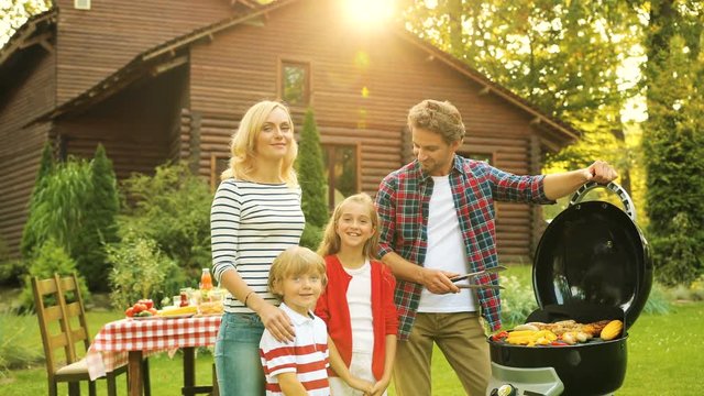 Happy parents with children near barbecue in the green yard with wooden house. Outdoors. Portrait