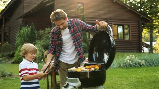 Attractive father grilling vegetables and sausages on the barbecue and his cute happy son helping him. Wooden cozy house on the backgrond. Sunny summer day. Outdoors