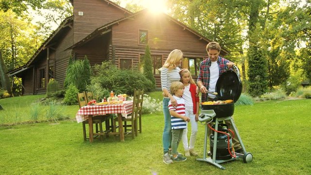 Good-looking father grilling vegetables and meat on the barbecue while his family looking at his work. Beautiful green yard, wooden cozy house and table with food on the background. Outside