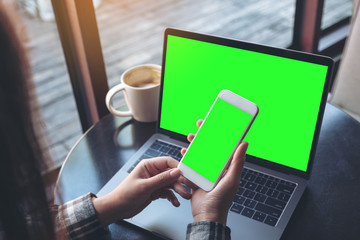 Mockup image of business woman holding mobile phone with blank green screen while using laptop on wooden table in cafe