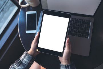 Top view mockup image of hands holding black tablet pc with white blank screen and laptop , coffee cup on wooden table background in cafe
