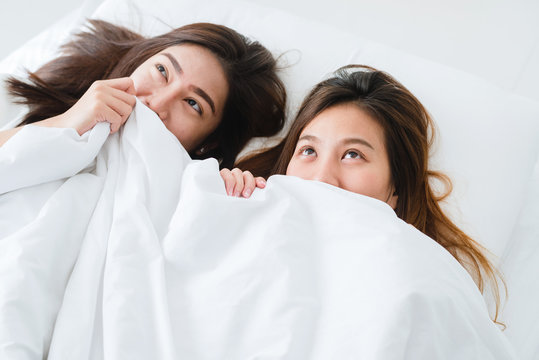 Top view of beautiful young asian women lesbian happy couple showing surprise and looking at camera while lying in bed under blanket. Funny women after wake up. Lesbian couple together indoors concept