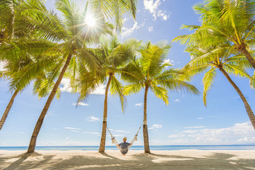 Tourist in hat sitting on hammock  on the beach between palms. Travel and vacation concept.