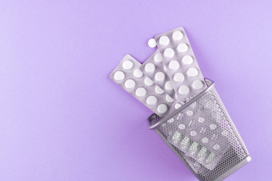 Trash bin with packs of white capsules and pills packed in blisters with copy space on purple background. Focus on foreground, soft bokeh