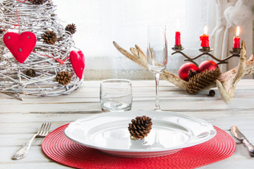 Christmas place setting with white dishware, silverware and red decorations on white board in interior near the window.